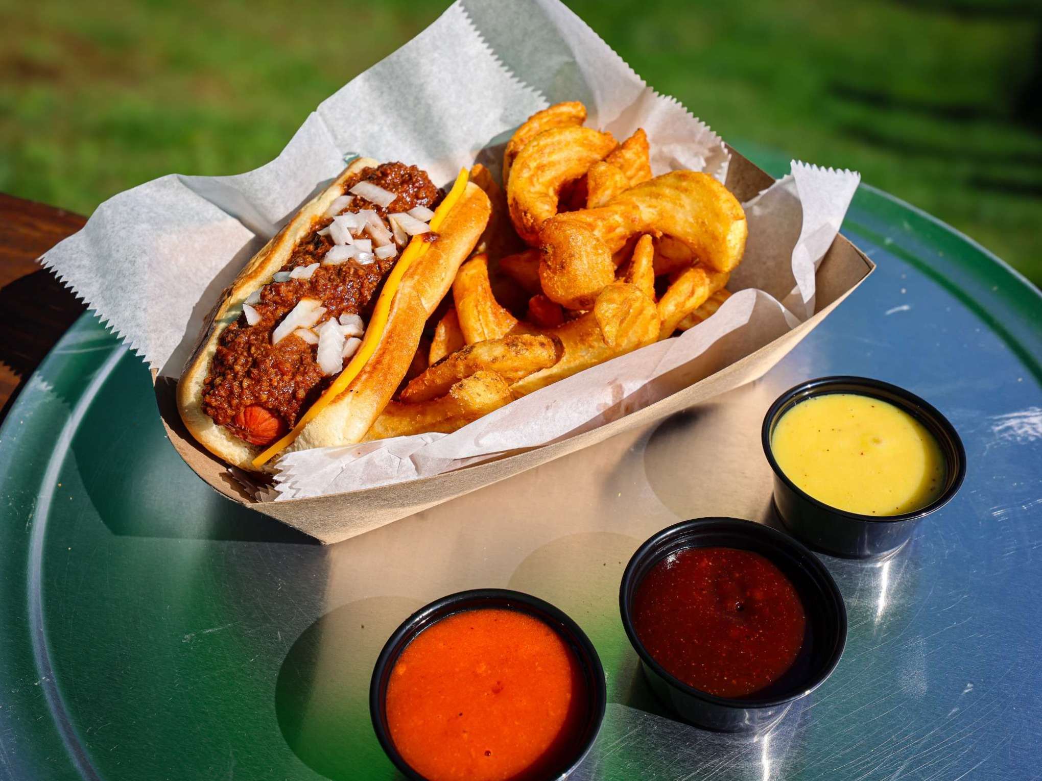 Hot dog with chili and onions, curly fries, and three dipping sauces on a metal tray.