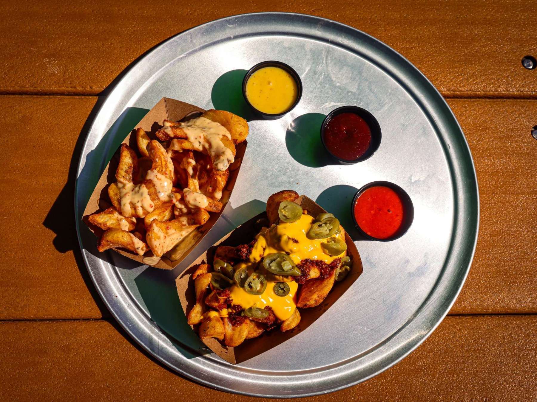 Two trays of loaded potato wedges with three sauce cups on a metal tray.