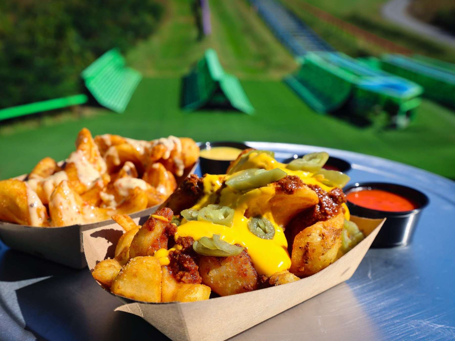 Loaded potato wedges with cheese and toppings on a tin table in front of a grassy hill.